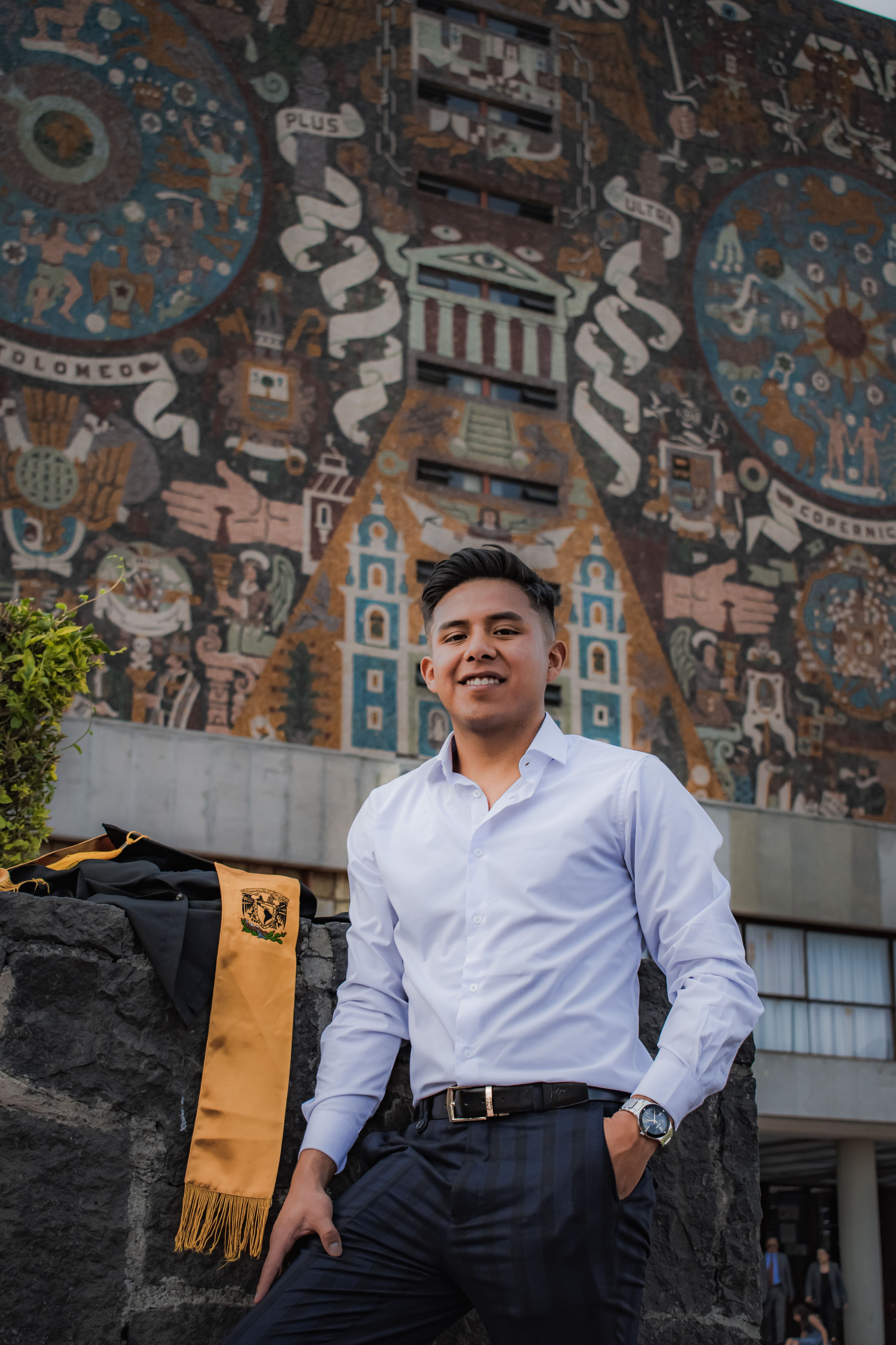 Young man in white shirt standing proudly in front of the iconic mosaic mural at UNAM, Mexico City.