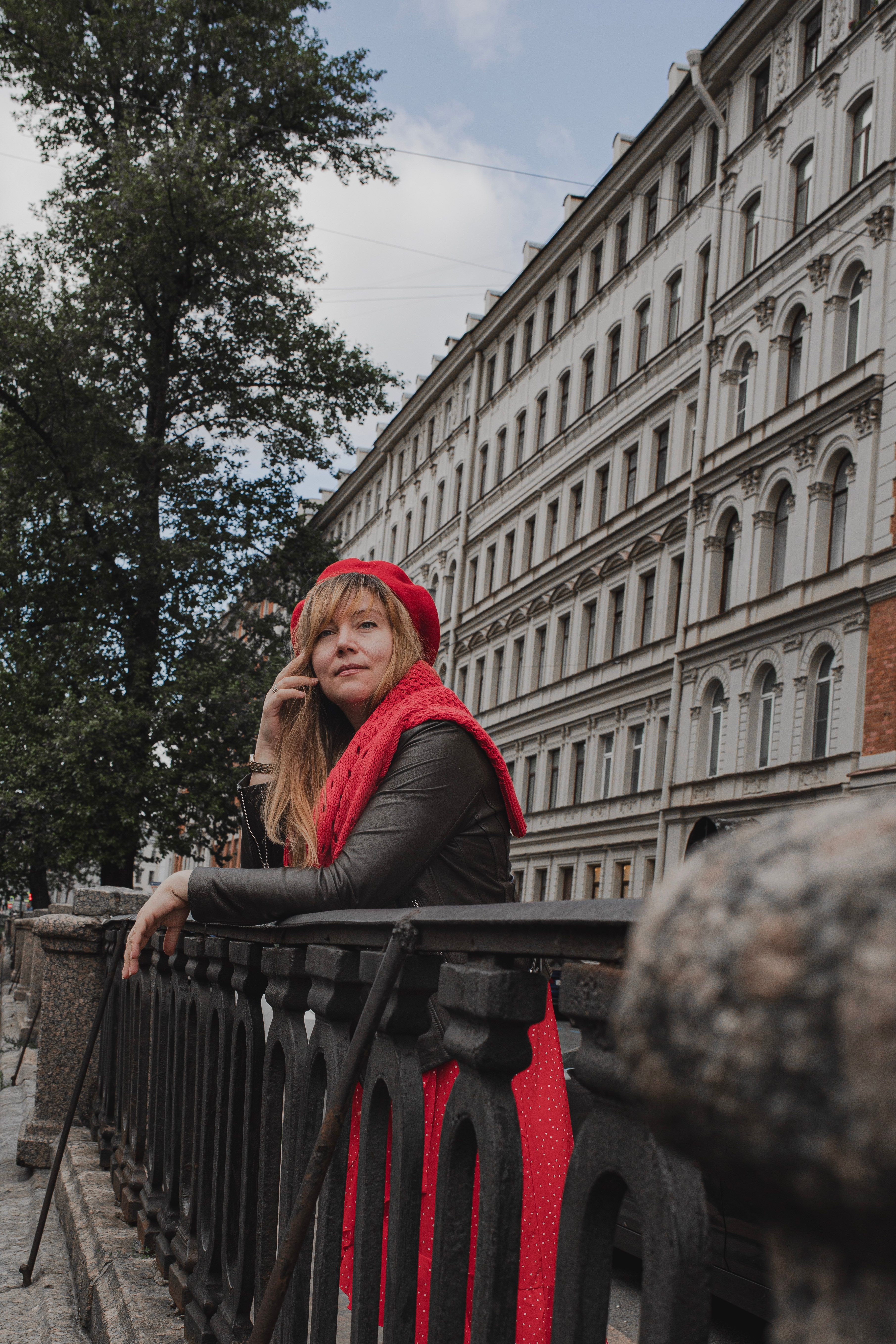 A fashionable woman wearing a red beret and scarf leans on a railing in an elegant city street.