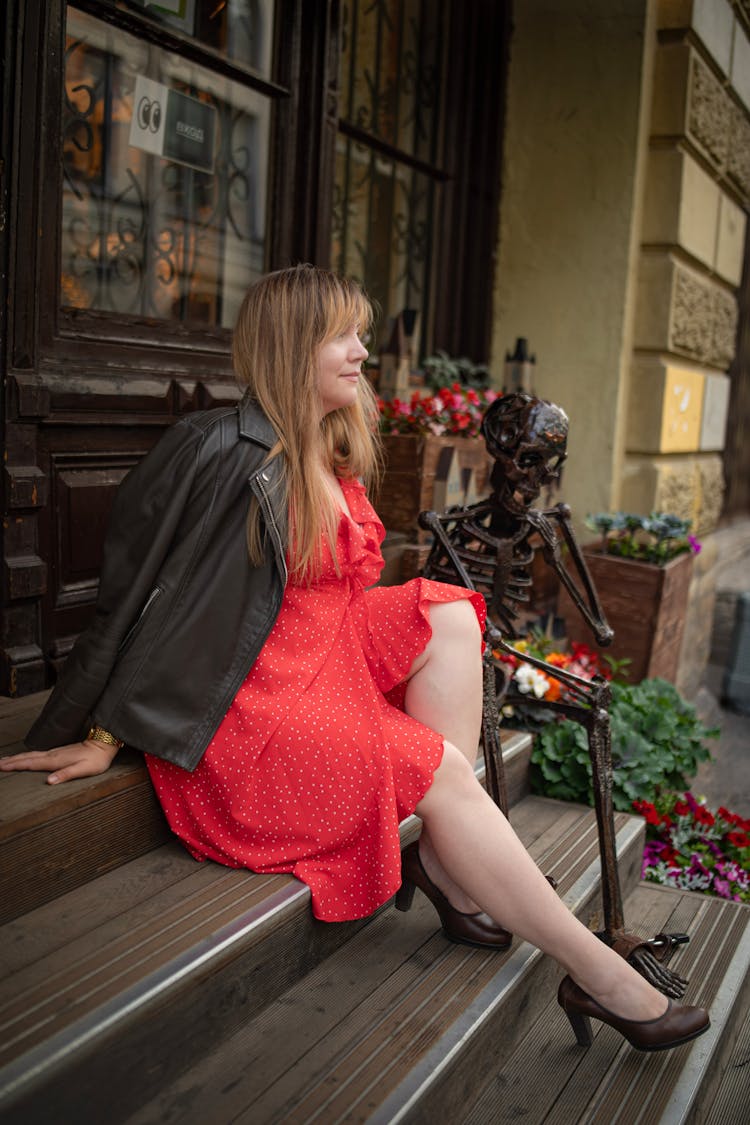 Woman In Black Leather Jacket And Red Dress Sitting On Steps Beside A Skeleton Sculpture