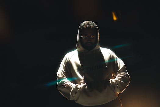 A man wearing a hoodie stands in dramatic lighting at night, hands in pockets, conveying a mysterious mood.