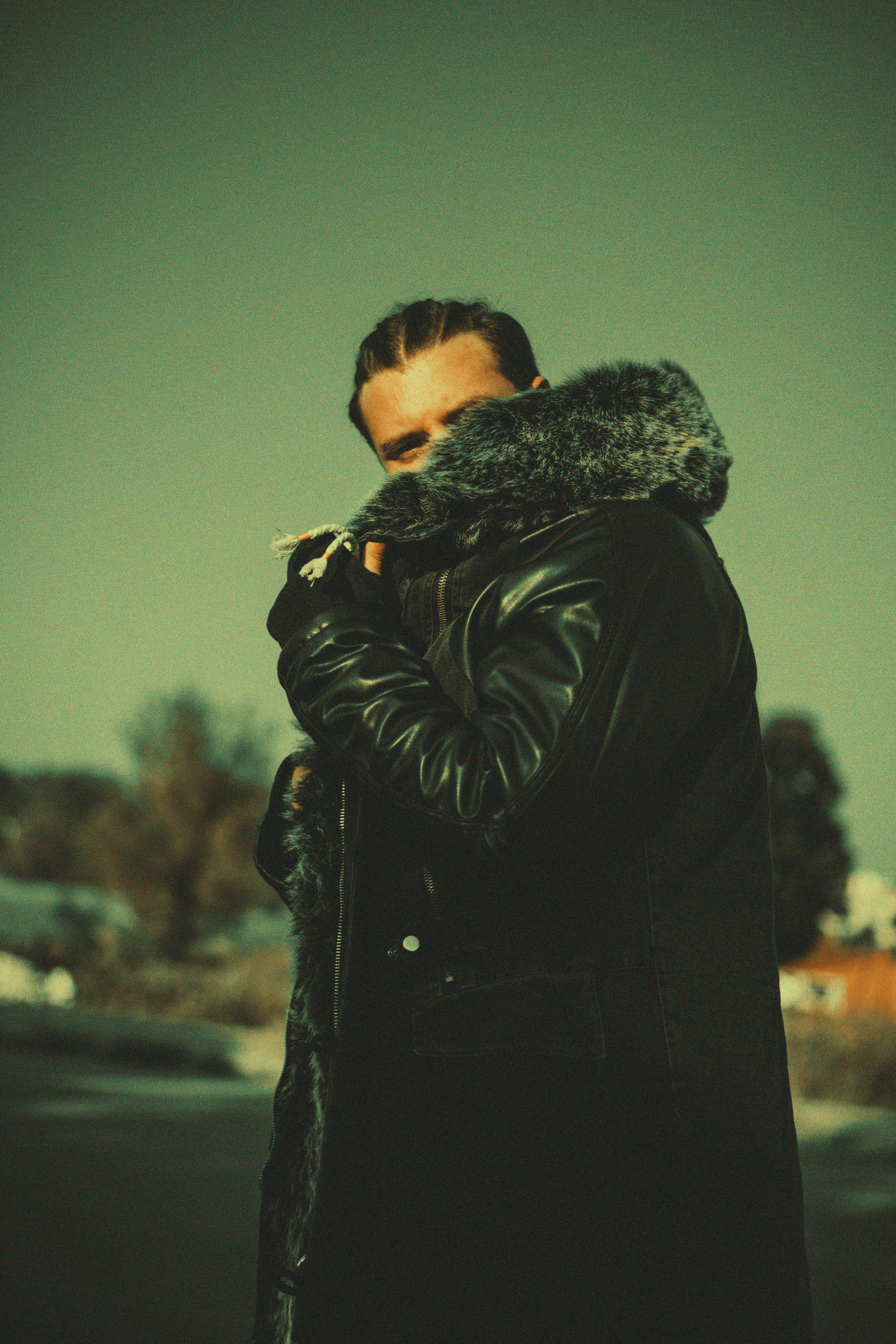 Man with braided hair wearing a leather coat with a fur collar standing outdoors on a cool day.