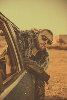 A man with sunglasses and fur coat leans out of a car window in a vintage setting.