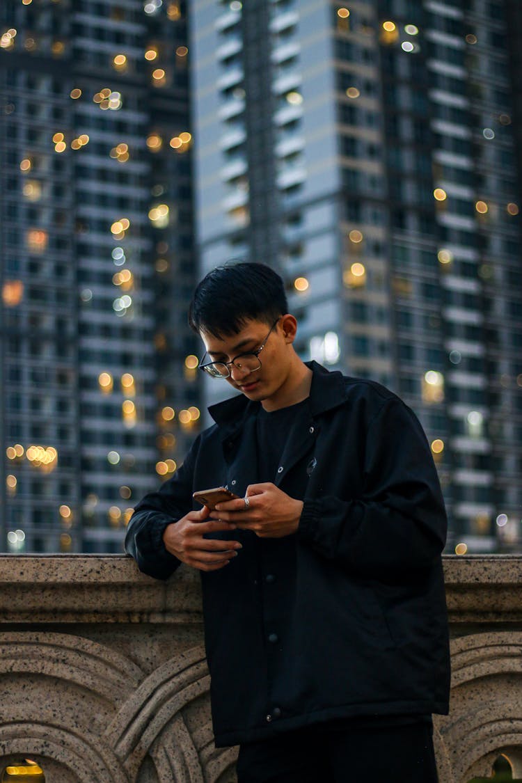 Young Man In Black Jacket Checking His Smart Phone On A City Street