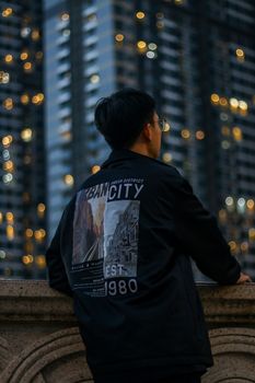 A man in a fashionable jacket poses against a cityscape with high-rise buildings in the evening.