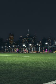 Nighttime soccer match with players under illuminated city skyline in an urban environment.