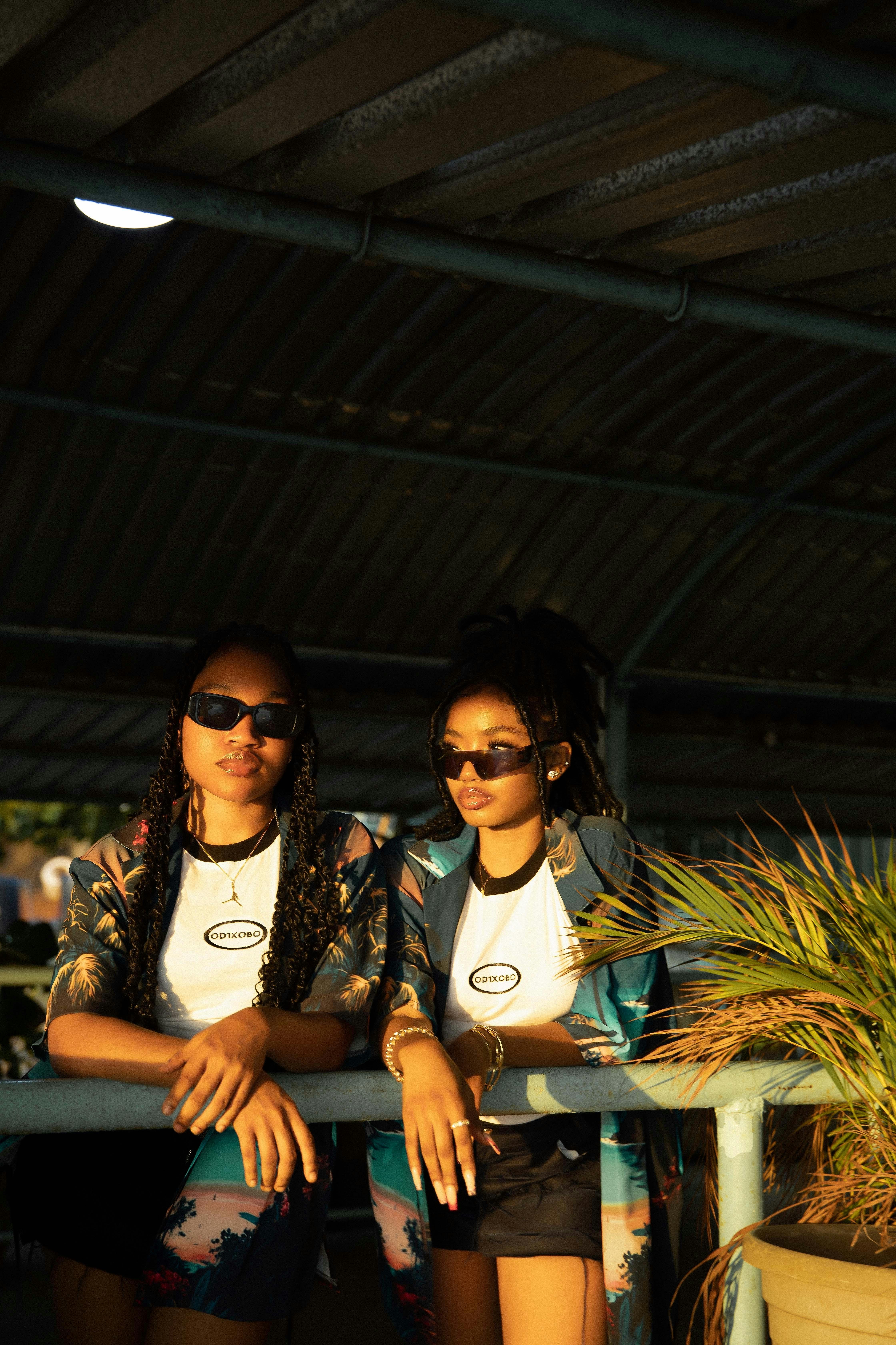 Two fashionable black women with sunglasses relax in a sunlit outdoor setting, showcasing modern casual style.