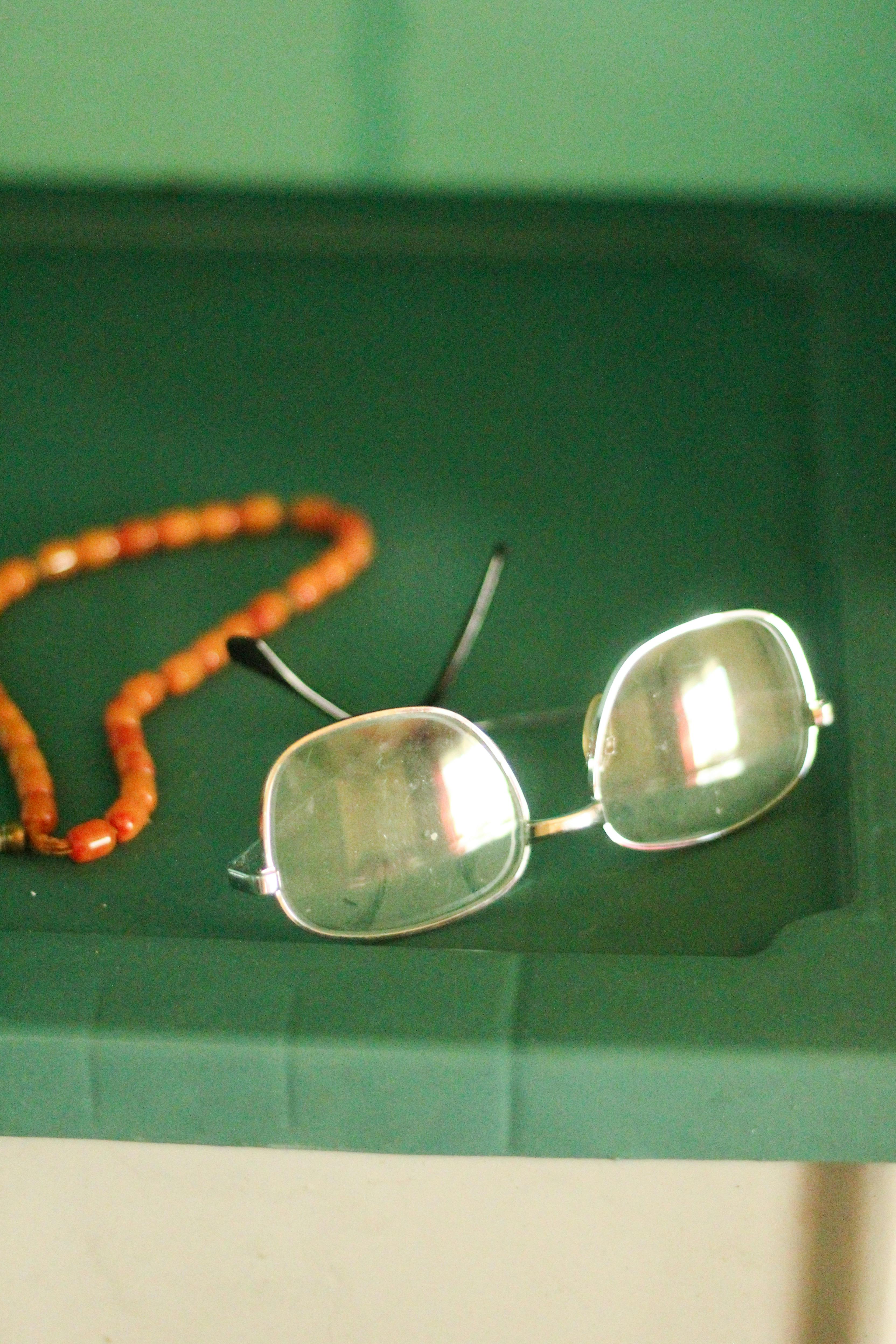 Close-up of eyeglasses and beaded necklace on green surface in a minimalist composition.