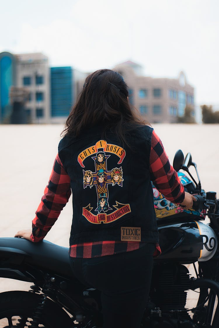 Biker In A Black Denim Vest With A Guns N Roses Print On The Back Posing By The Motorcycle