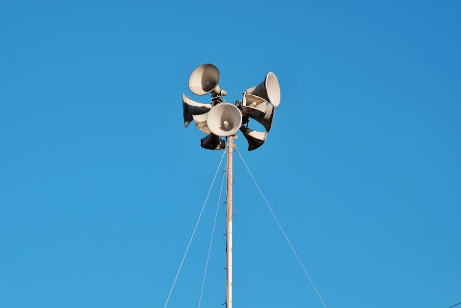 High pole with multiple loudspeakers against a vibrant blue sky, urban setting.