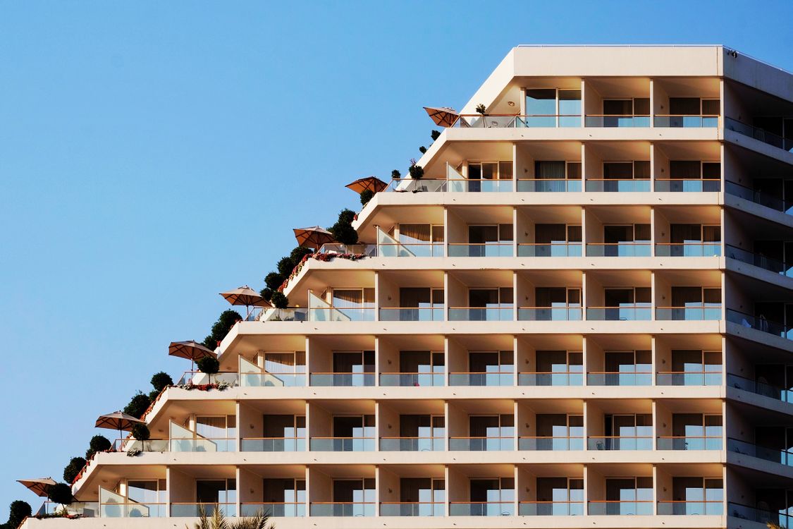 Modern terraced resort hotel exterior with balconies, umbrellas, and greenery against a clear blue sky