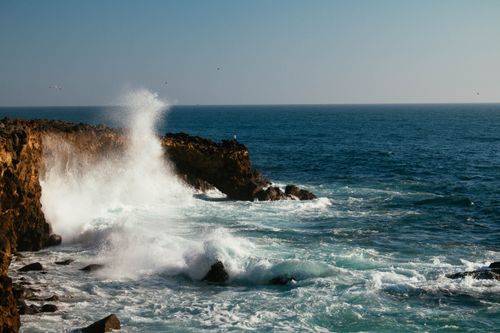 Free Powerful ocean waves crash against a rugged cliff under a clear blue sky, capturing coastal beauty and energy. Stock Photo