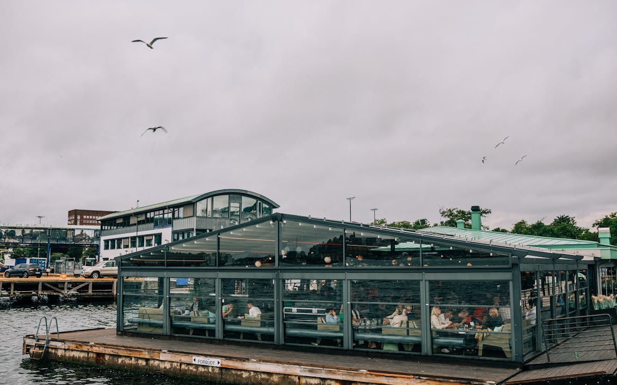 People enjoying a meal in a modern glass restaurant by the harbor on a cloudy day.