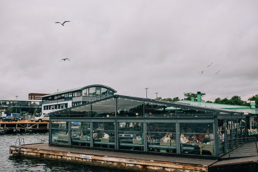 People enjoying a meal in a modern glass restaurant by the harbor on a cloudy day.
