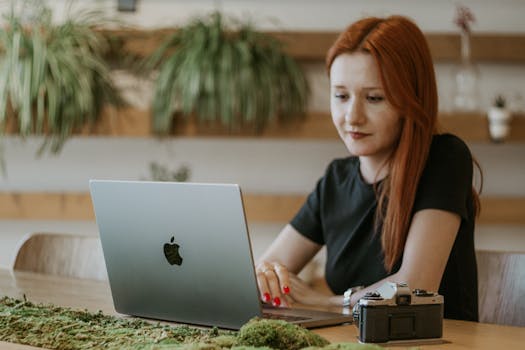 A focused red-haired woman working on a laptop in a modern indoor setting.