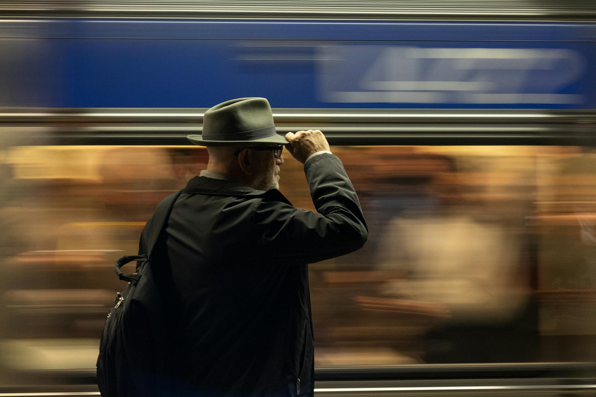 An elderly man in a hat stands on a subway platform with a train speeding by, capturing urban life.