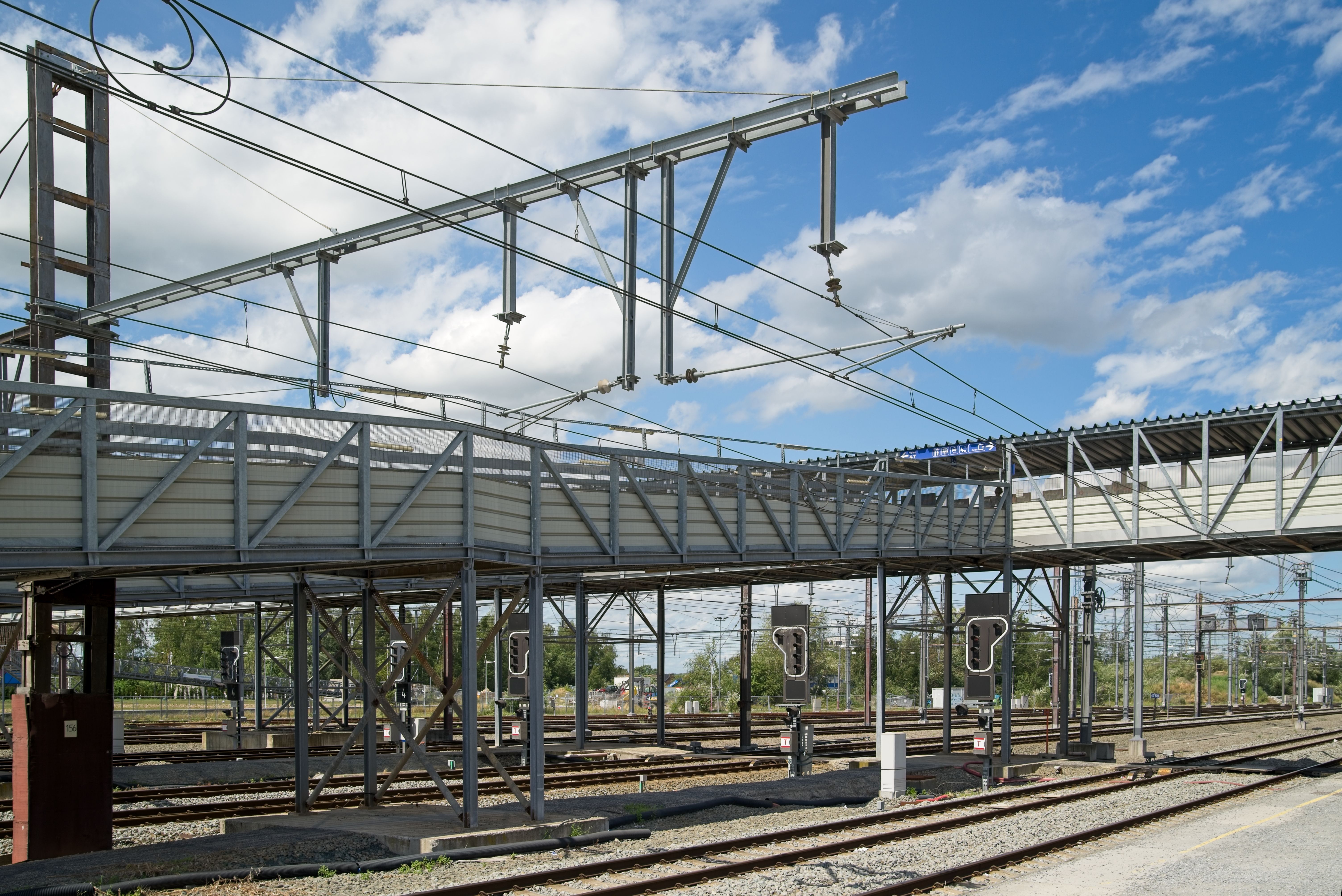 A contemporary railway station with overhead wires under a bright blue sky with clouds.