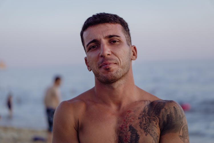 Shirtless Brunette Man With Shoulder Tattoos Posing On A Beach