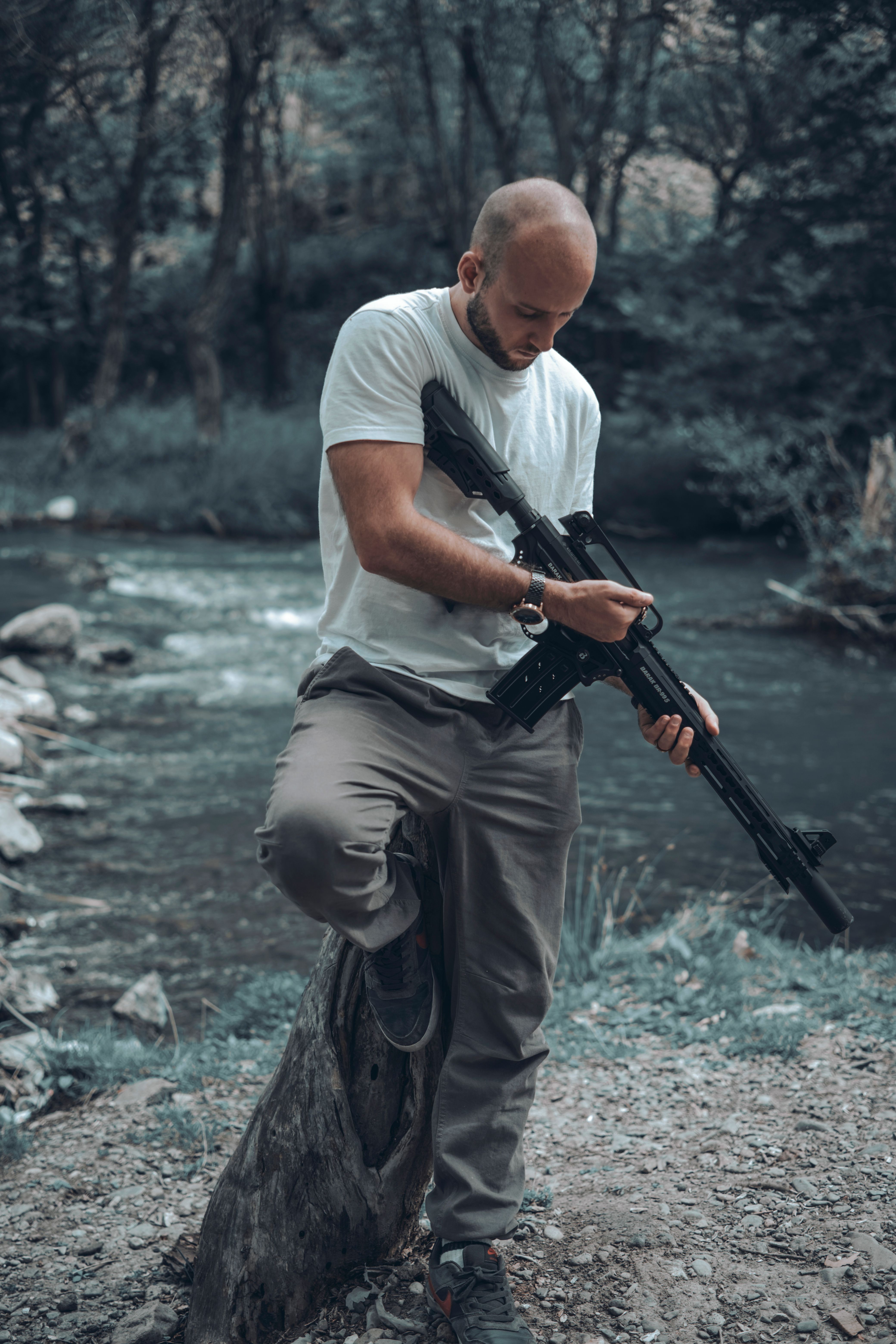 A man standing by a forest stream holding a rifle, creating a contemplative outdoor moment.