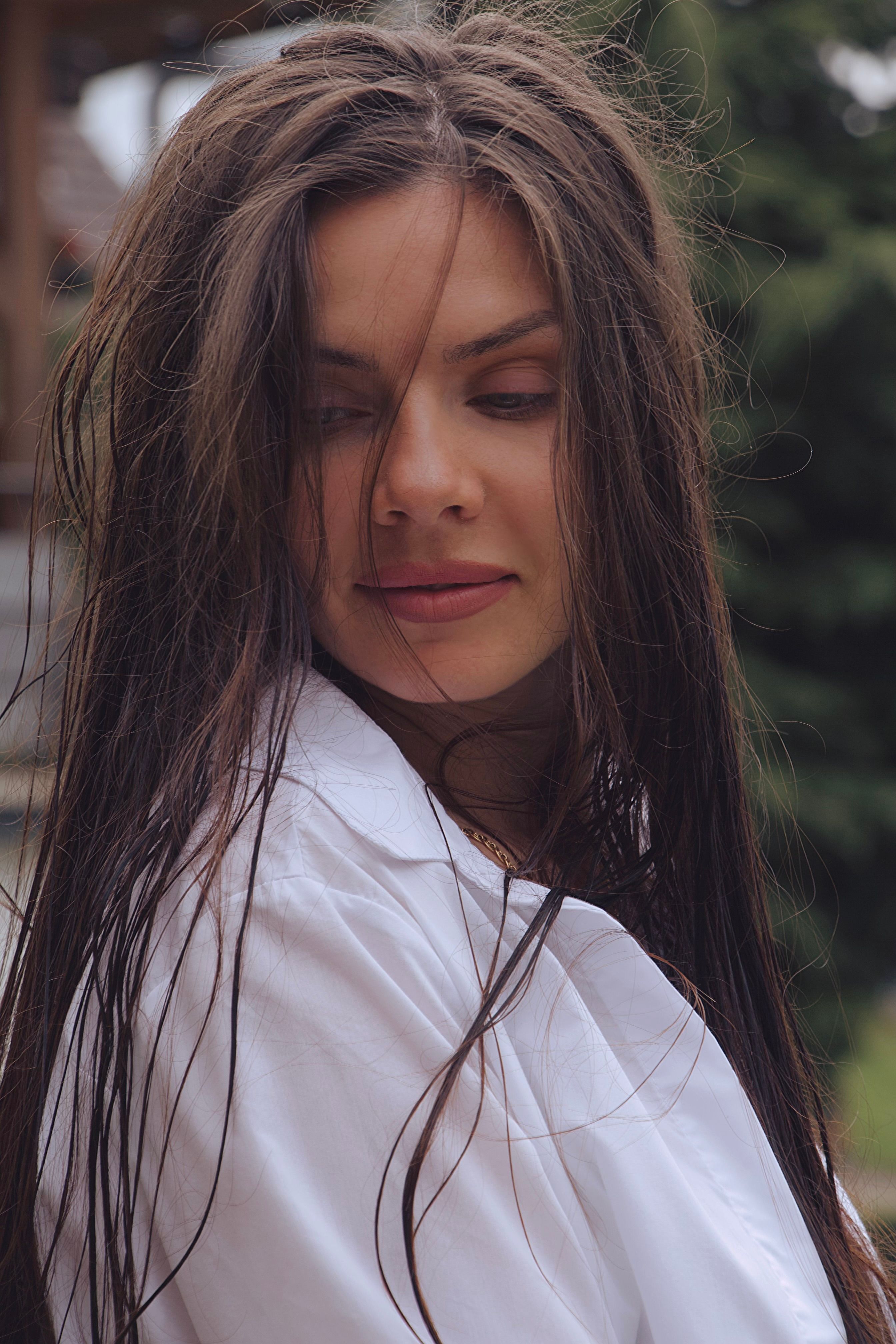 Close-up of a woman with tousled hair in a white shirt, looking down outdoors.