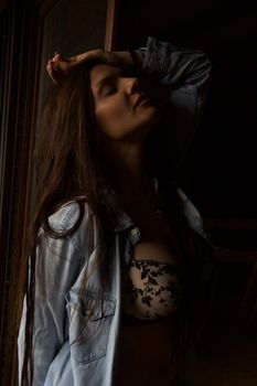 Stylish woman in a dark setting, wearing a denim shirt and floral lingerie, posing confidently.