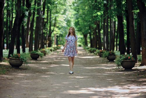 Free A woman strolls along a tree-lined path with potted flowers in a forest during summer. Stock Photo
