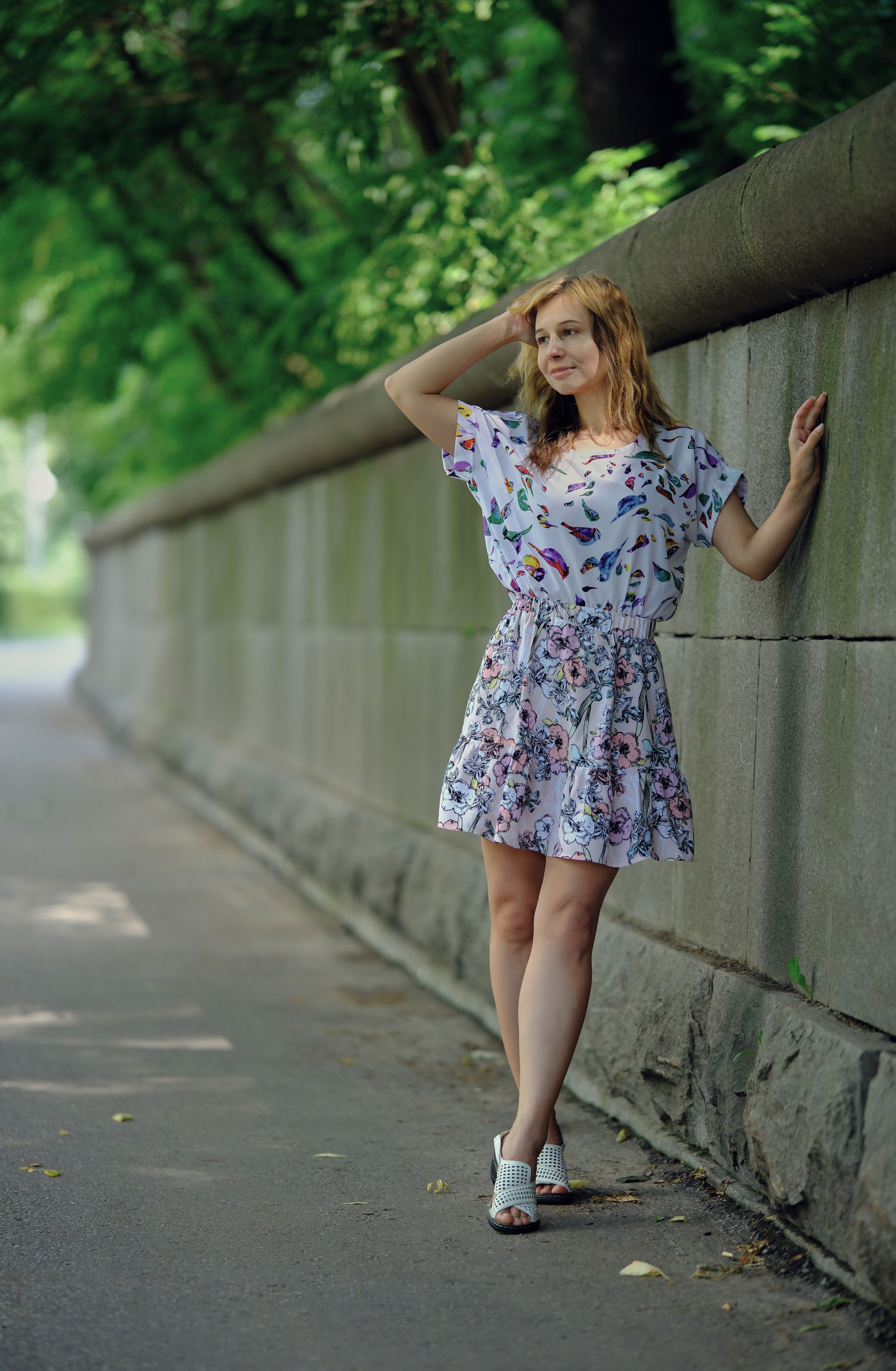 Stylish woman in floral dress leans against a stone wall on a tree-lined sidewalk.