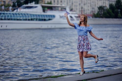 Free A woman stands by a river with a paper plane, posing in an urban environment with a ship in the background. Stock Photo