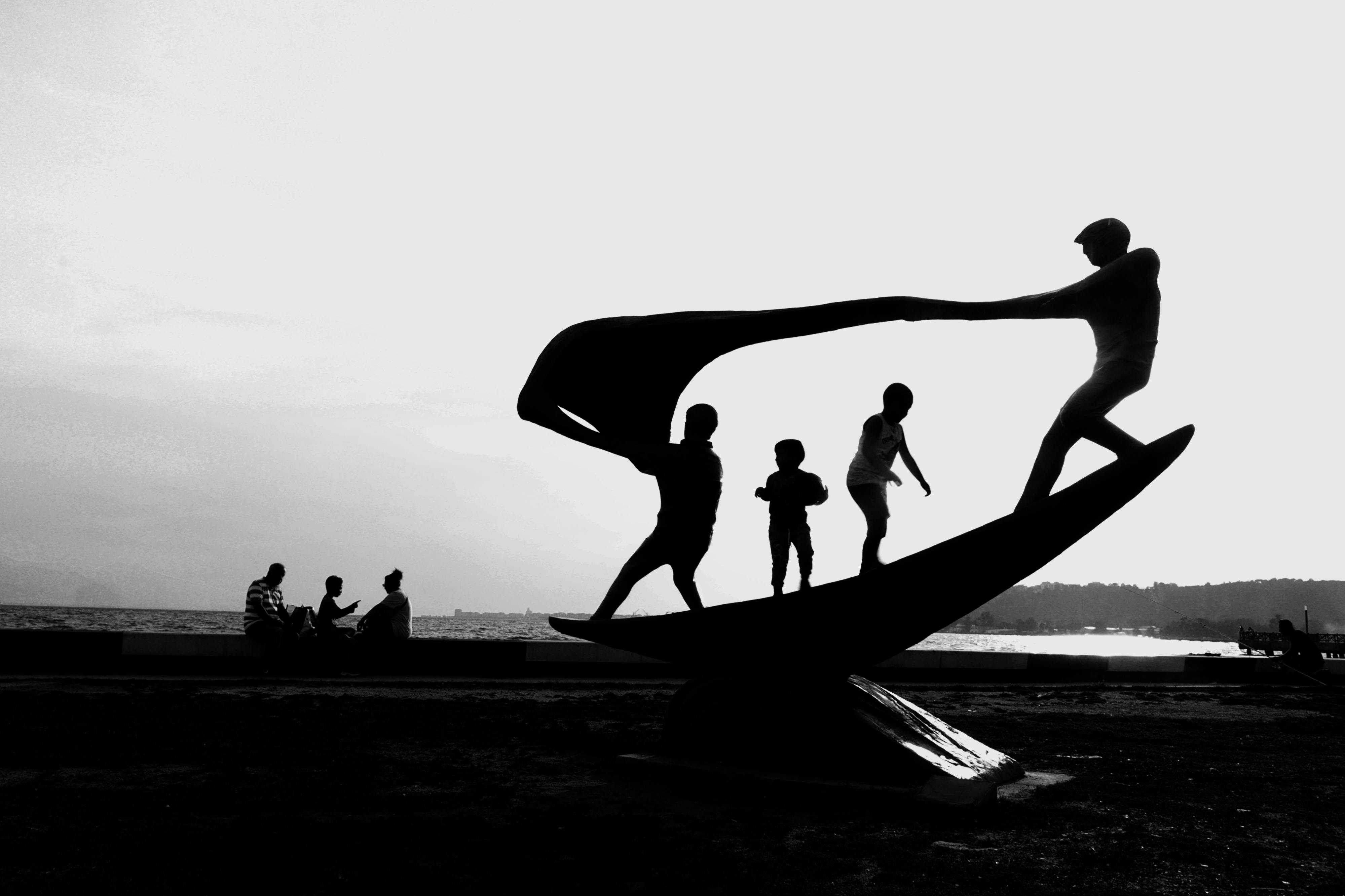 Black and white silhouette of children and adults at a seaside sculpture during sunset.