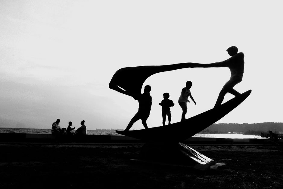 Black and white silhouette of children and adults at a seaside sculpture during sunset.