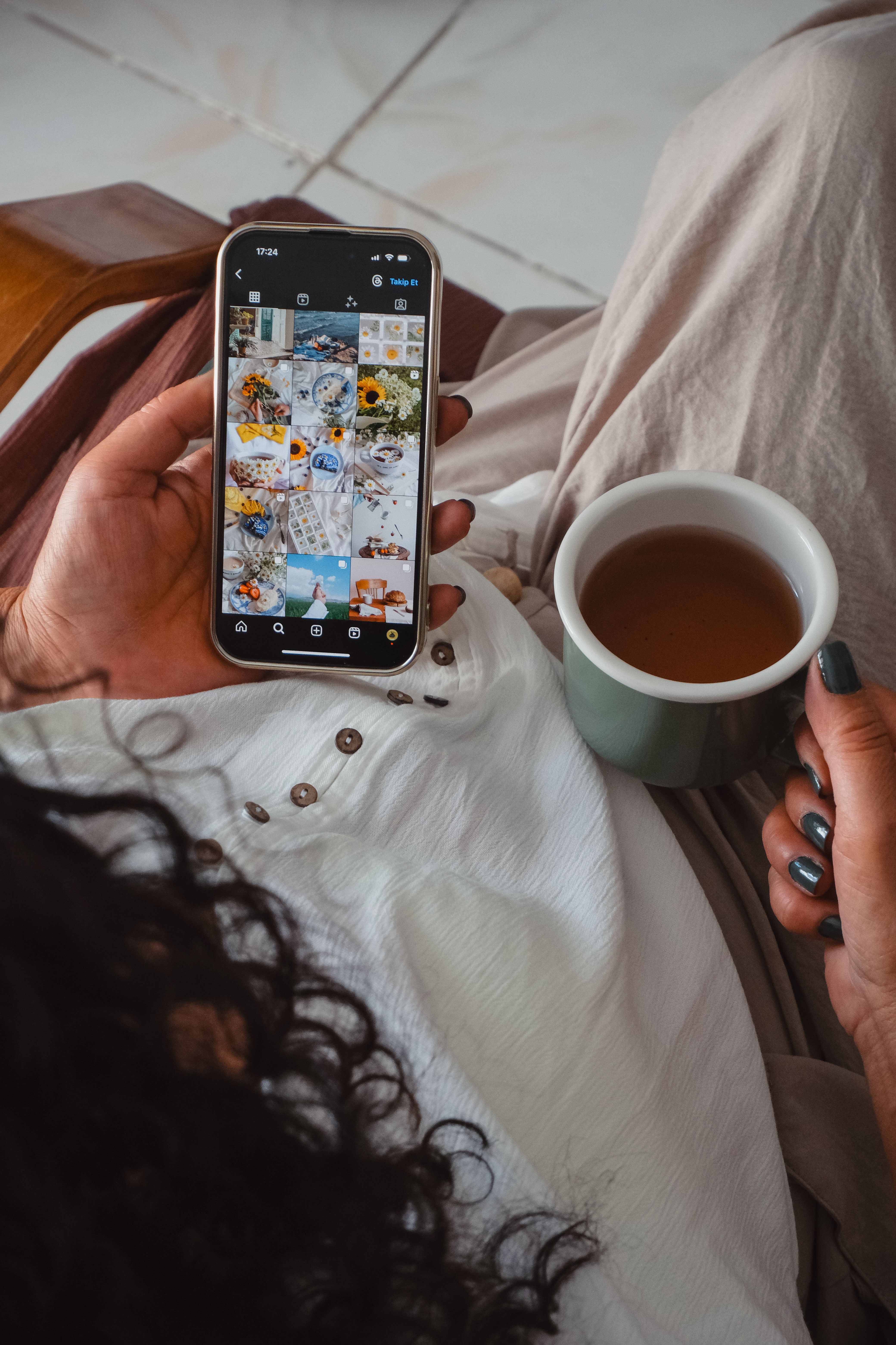 Woman holding smartphone and cup of tea, browsing images in a cozy indoor setting.