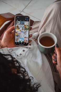Woman holding smartphone and cup of tea, browsing images in a cozy indoor setting.