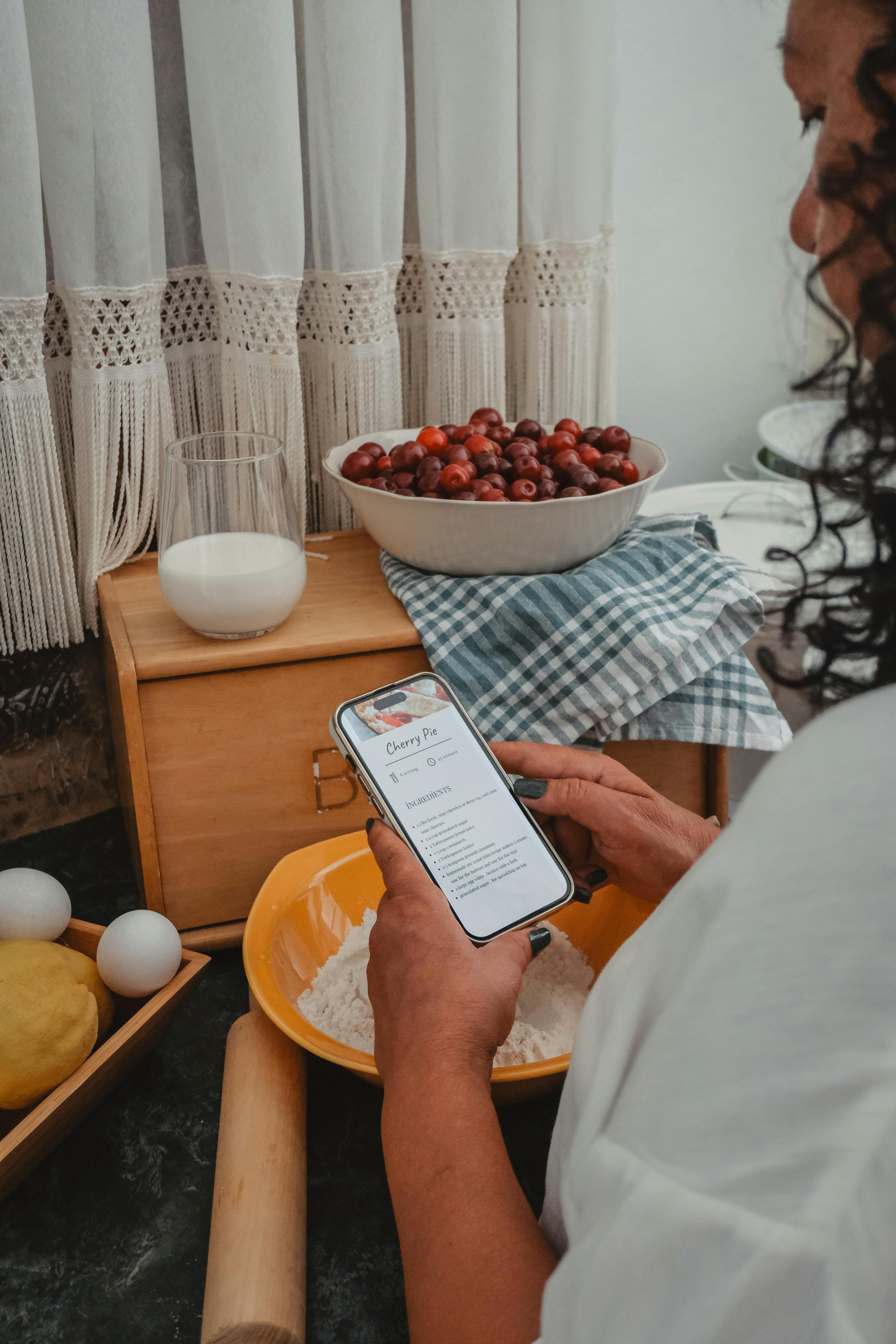 A woman follows a cherry pie recipe on her phone, surrounded by ingredients in her kitchen.