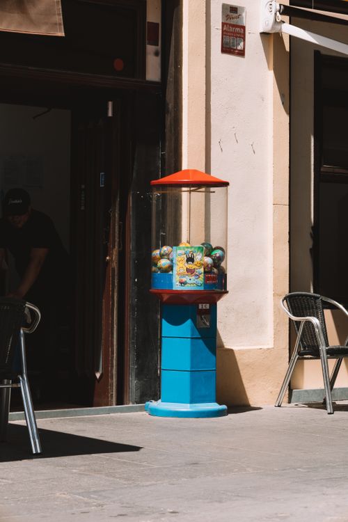 Free A toy vending capsule on a sunlit sidewalk with nearby chairs and urban elements. Stock Photo
