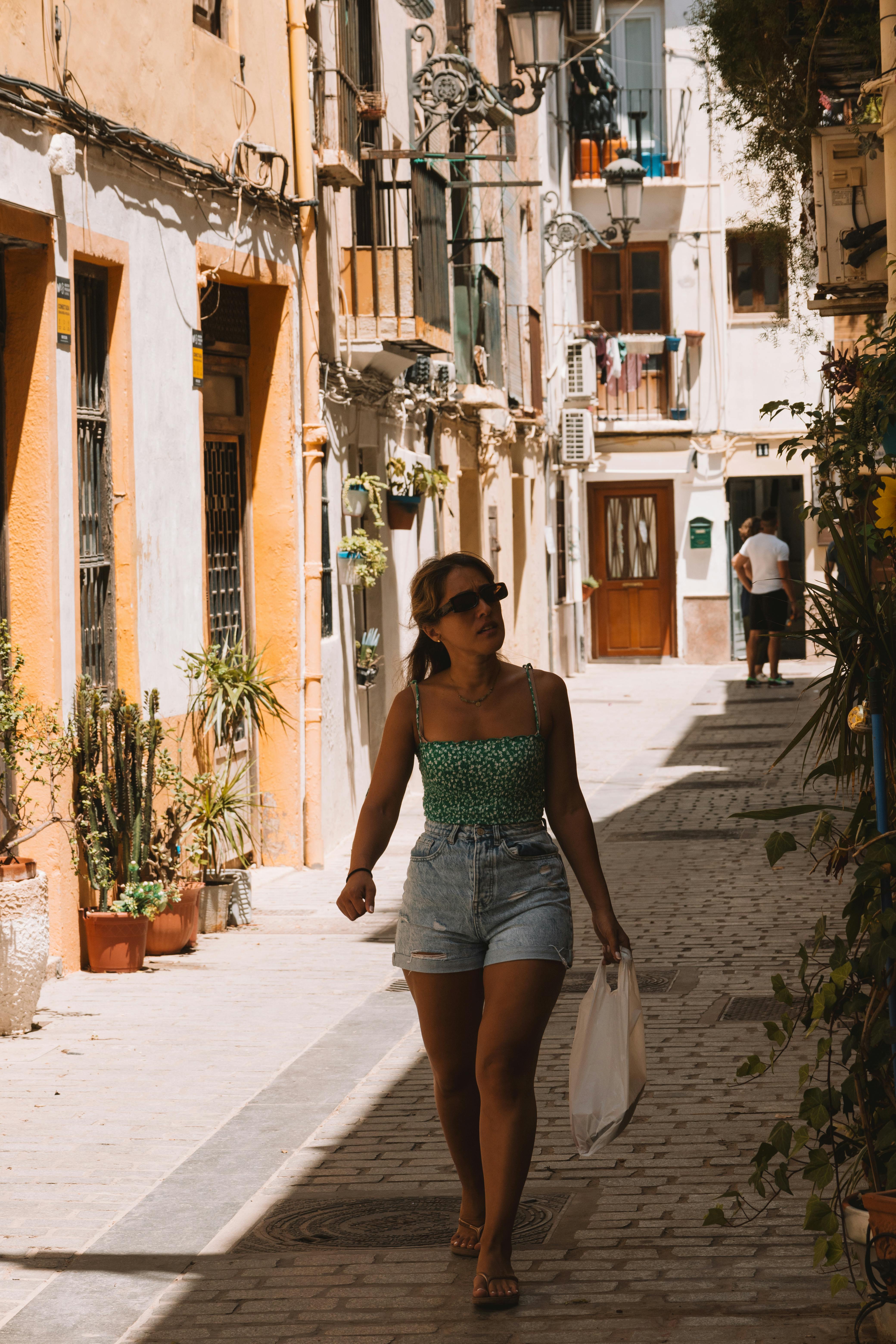 Free A woman walks through a sunlit, narrow street lined with potted plants and classic architecture. Stock Photo