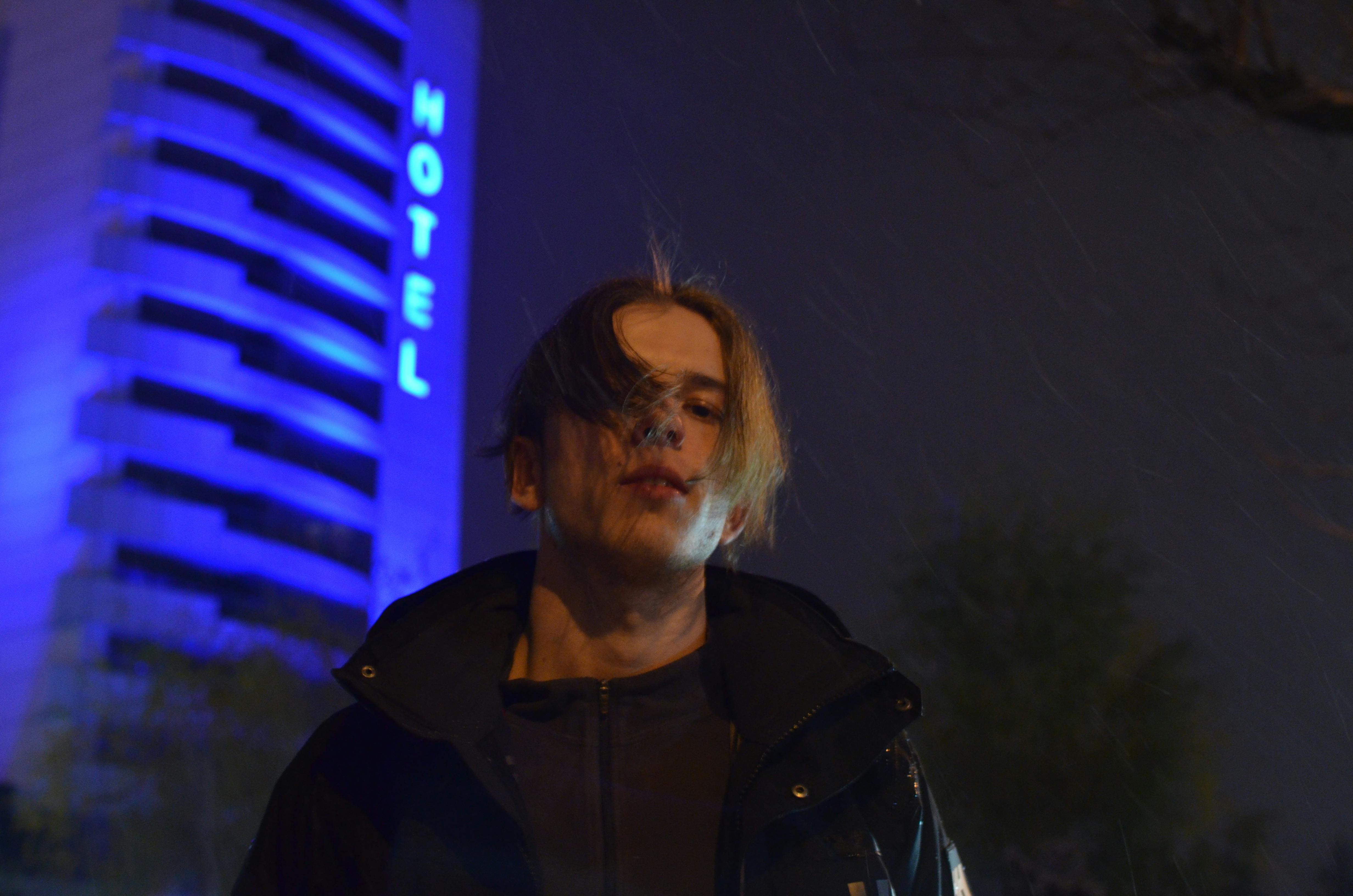 A moody portrait of a man in front of a hotel, illuminated by blue lights at night.
