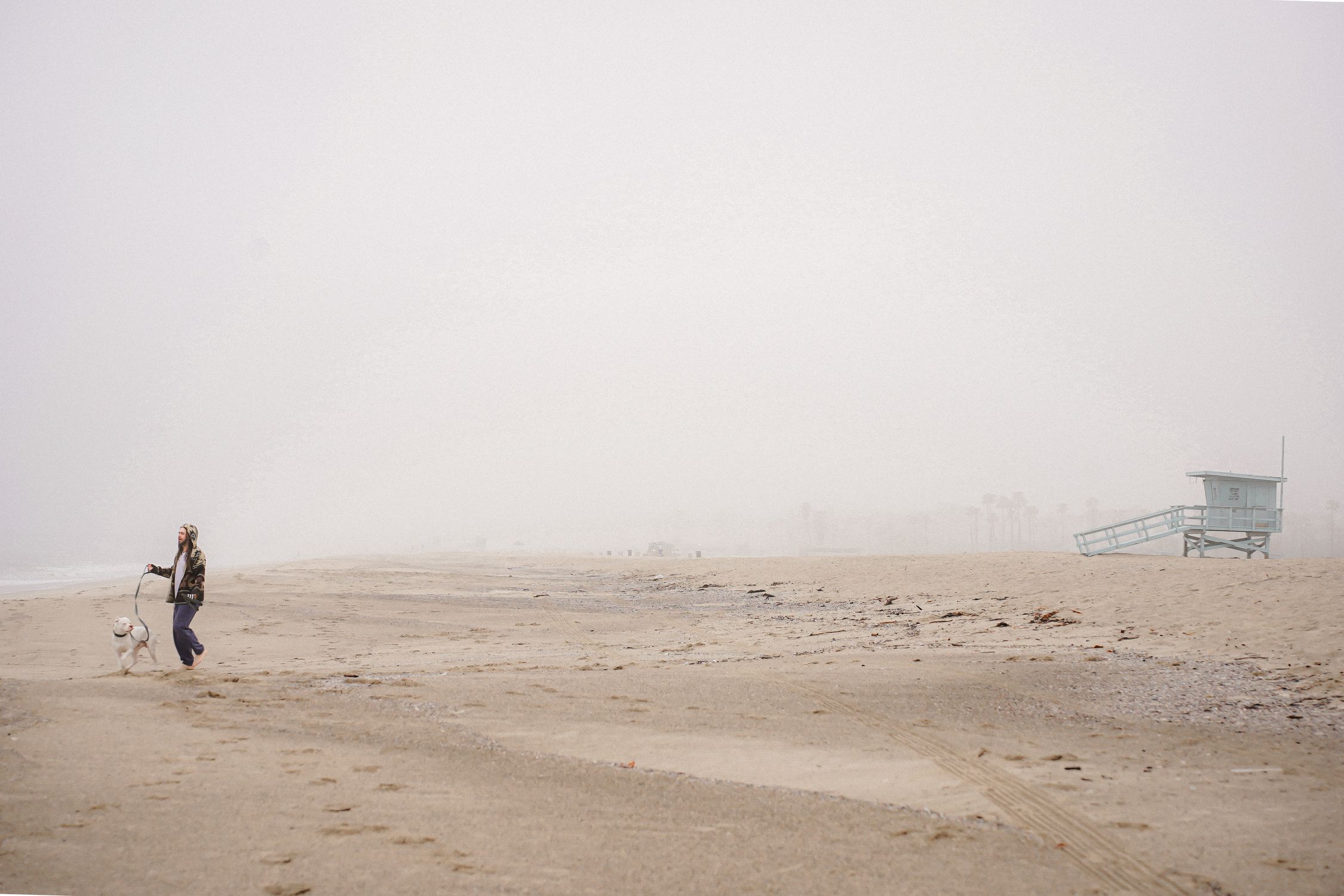 A woman strolling her dog on a foggy beach next to a lifeguard tower.