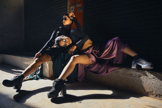 Two women in elegant dresses and sunglasses pose stylishly on a sunny day.