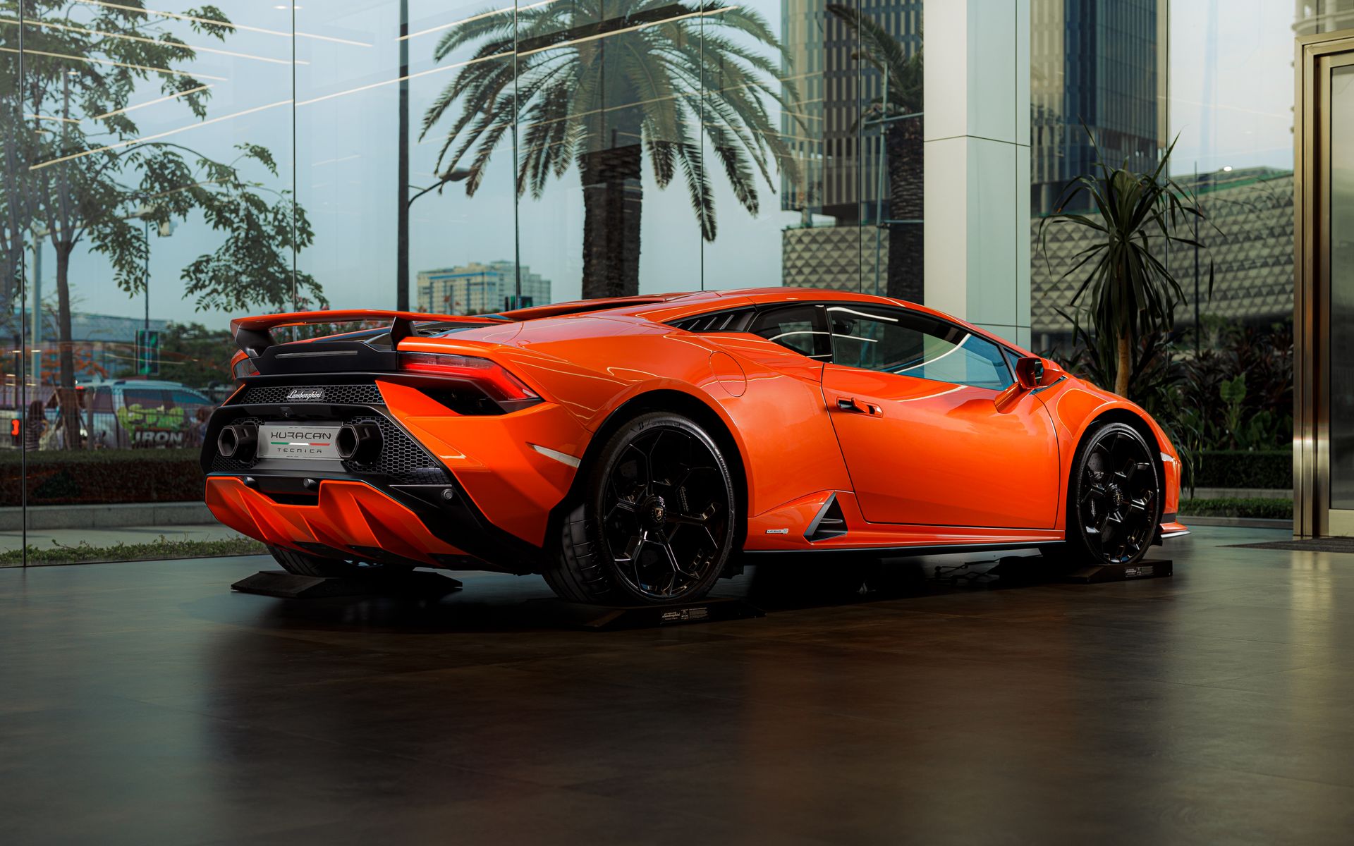 Bright orange sports car displayed in a sleek, modern dealership setting with cityscape in the background.