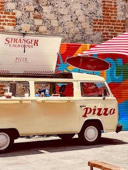 Retro pizza van parked by brick wall mural in a city street scene.