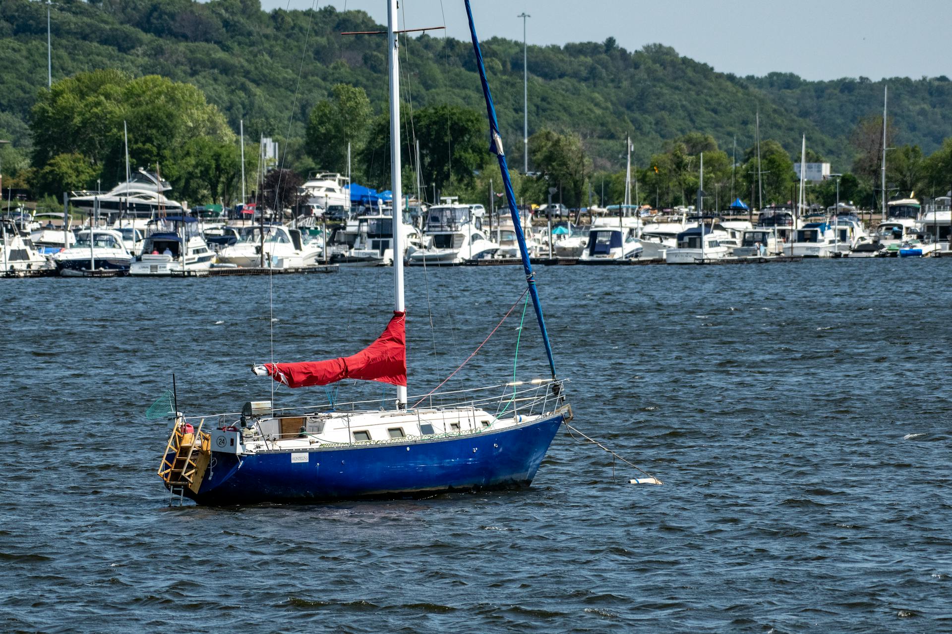 A sailboat anchored on the St. Croix River, Hudson, WI, with lush greenery and marina in the background.