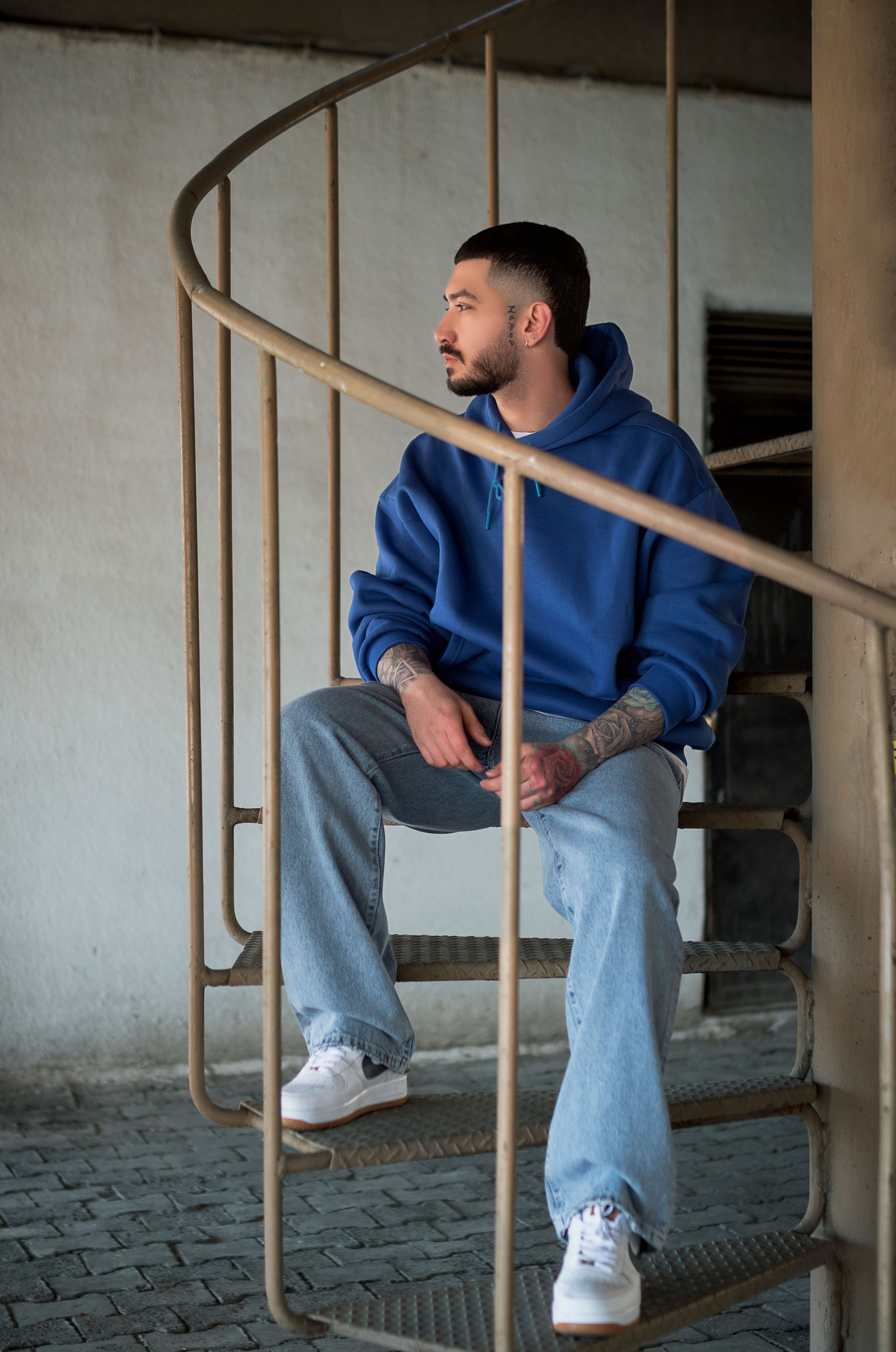 Man in blue hoodie sitting on a spiral staircase indoors, casual style.