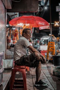 A man sitting in Bangkok's vibrant night market, captured in a moody and atmospheric setting.