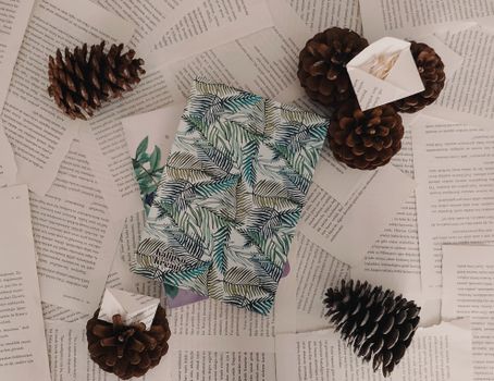 A beautifully wrapped present with leaf patterns surrounded by scattered book pages and pine cones.