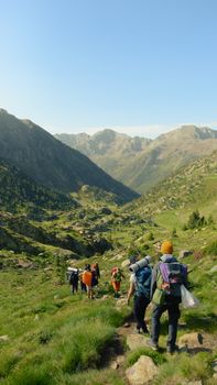 A group of hikers descends a lush mountain trail surrounded by stunning valleys under a clear blue sky.