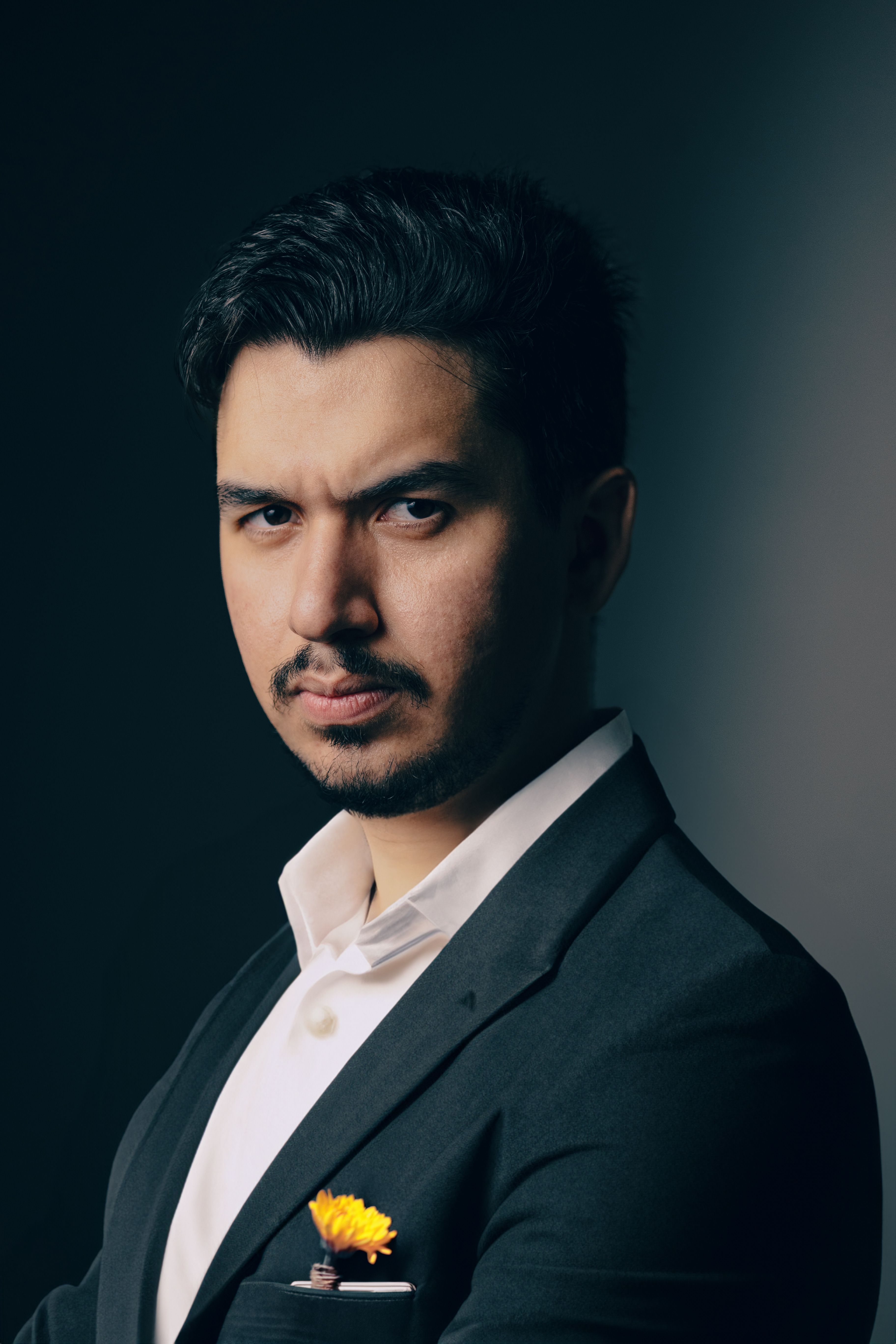 Portrait of a confident man in a black suit with a yellow flower, set against a dark background.