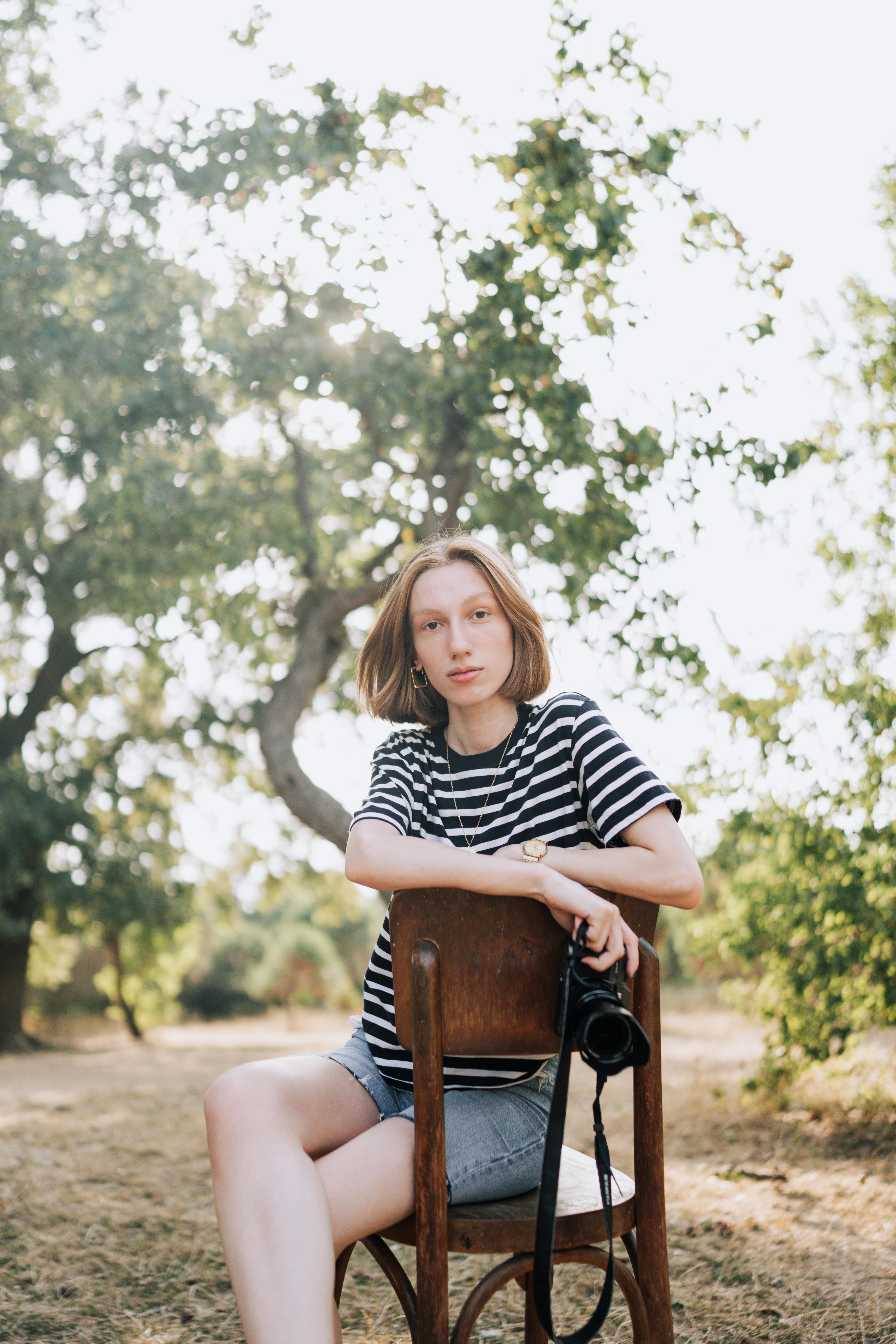 A young woman sits on a chair outdoors in a summer setting, holding a camera and looking relaxed.