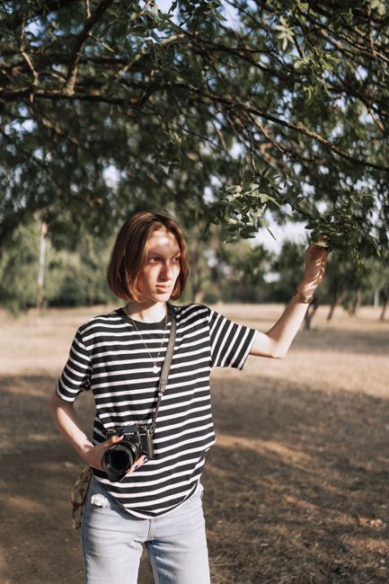 A young woman in a striped t-shirt holds a camera in a summer countryside setting.