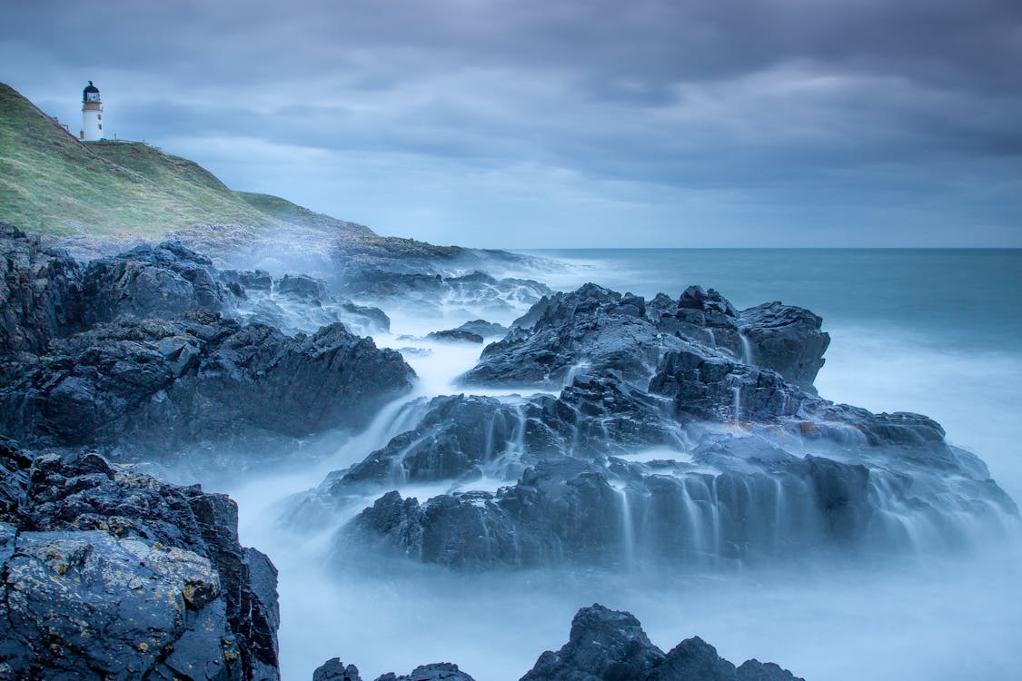 Free Captivating seascape of a lighthouse and rugged coastline with waves crashing against the rocks in Scotland. Stock Photo