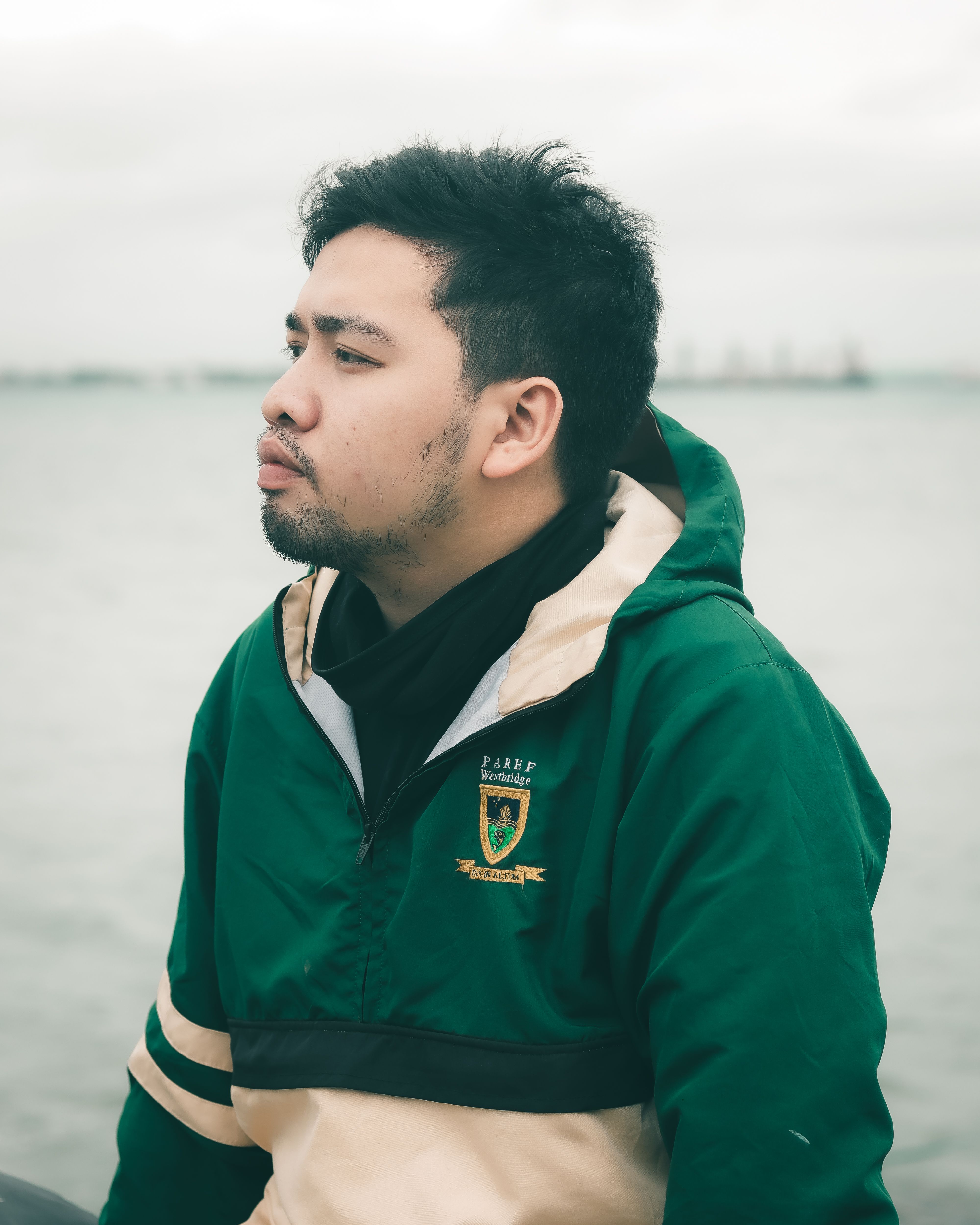 A man in a green jacket sits pensively by the sea, reflecting on the day.