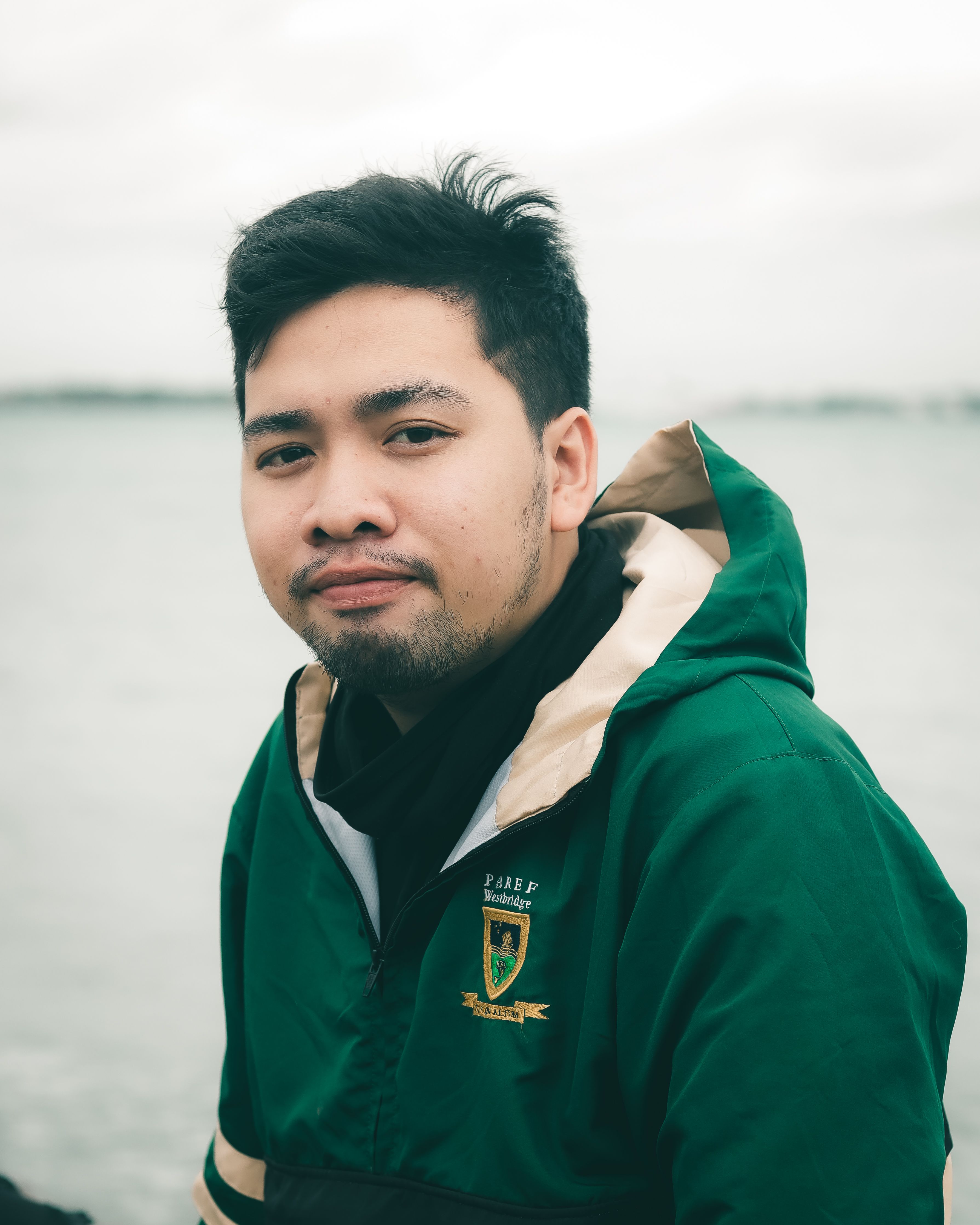 Young man in a green anorak sitting by the sea, looking content. Fashion photography outdoors.